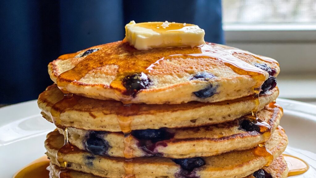 A tall stack of golden-brown blueberry cornmeal pancakes, generously drizzled with real maple syrup and topped with fresh blueberries and a pat of butter, set on a white plate.