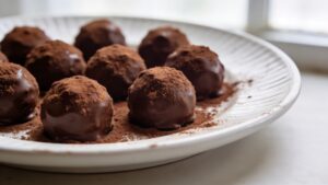 A close-up photo of rich, dark chocolate truffles dusted with cocoa powder, nuts, and coconut, arranged on a rustic wooden board
