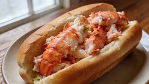 A close-up of a classic New England lobster roll on a plate, showing the split-top bun overflowing with tender lobster meat, with lemon wedge and side garnish