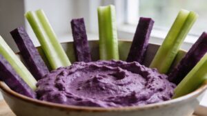 Colorful vegetable lightsabers arranged around a bowl of vibrant purple hummus on a dark background