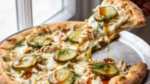 Whole dill pickle pizza with creamy garlic sauce, shredded chicken, mozzarella cheese, sliced pickles, hot honey drizzle, and fresh dill on a baking tray
