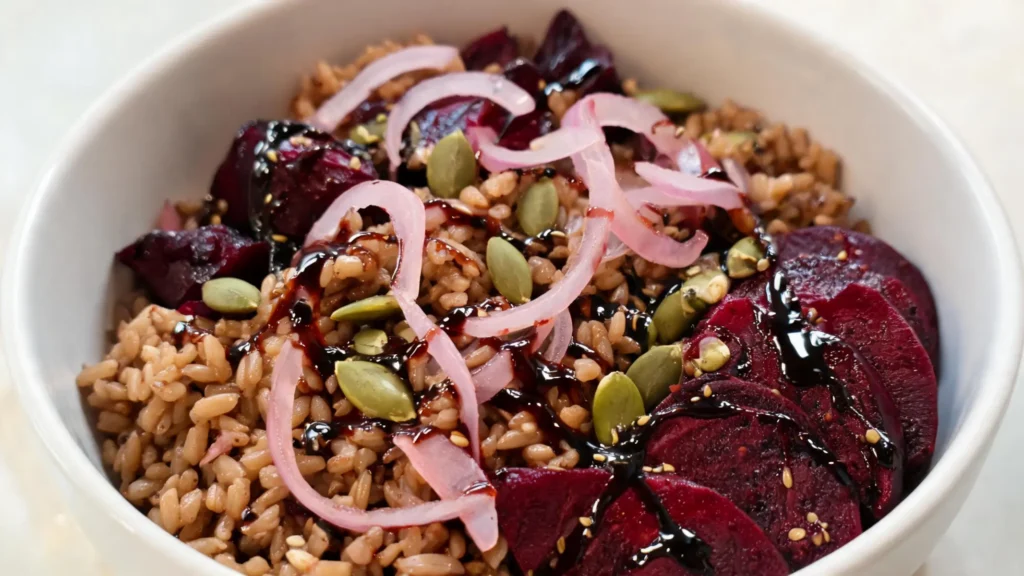A vibrant grain bowl with roasted beets, farro, pickled shallots, pepitas, and balsamic drizzle on a wooden table.