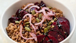 A vibrant grain bowl with roasted beets, farro, pickled shallots, pepitas, and balsamic drizzle on a wooden table.