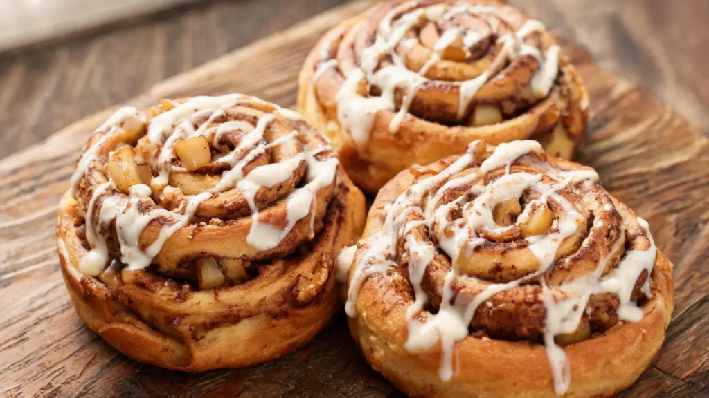 Freshly baked apple cider cinnamon rolls topped with white glaze on a wooden board with autumn leaves