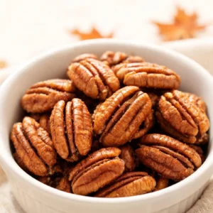 A bowl of pumpkin spice candied pecans with cinnamon sticks and autumn leaves